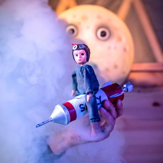 Billede af en dukke, der sidder på en raket. Billede fra forestillingen.