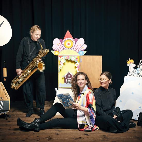 tre musikere, en med en saxofon, en sidder med bogen Skrædderkaninerne og bingebjørnen rejser i tiden.