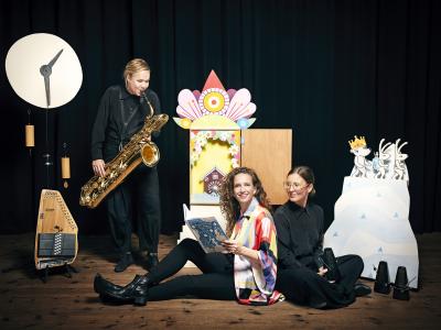 tre musikere, en med en saxofon, en sidder med bogen Skrædderkaninerne og bingebjørnen rejser i tiden.