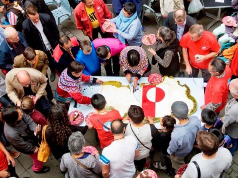 En masse mennesker i grønlandske nationaldragter samlet om et bord med det grønlandske flag.