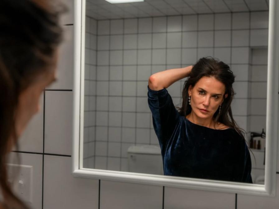 Billede fra filmen The Substance, hvor Demi Moore står foran et spejl og roder sig i håret.