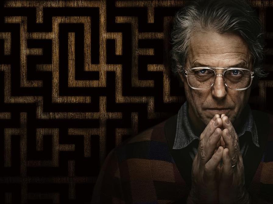 Billede af skuespilleren Hugh Grants ansigt på en mørk baggrund med et mønster. Han folder hænderne foran ansigtet.